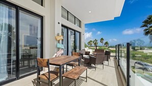 Un patio moderno al aire libre con una mesa y sillas de madera, ventanas amplias y vistas a palmeras y una carretera.