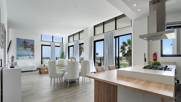 Una cocina moderna con una isla central, ventanas amplias y un comedor con sillas blancas.