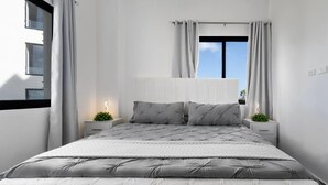 Un dormitorio con una cama, dos mesitas de noche, una ventana con cortinas y vistas a edificios.