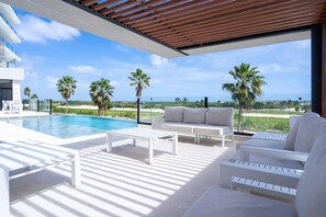 Un espace de vie extérieur moderne, doté d’une piscine, de mobilier blanc et offrant une vue sur l’océan.