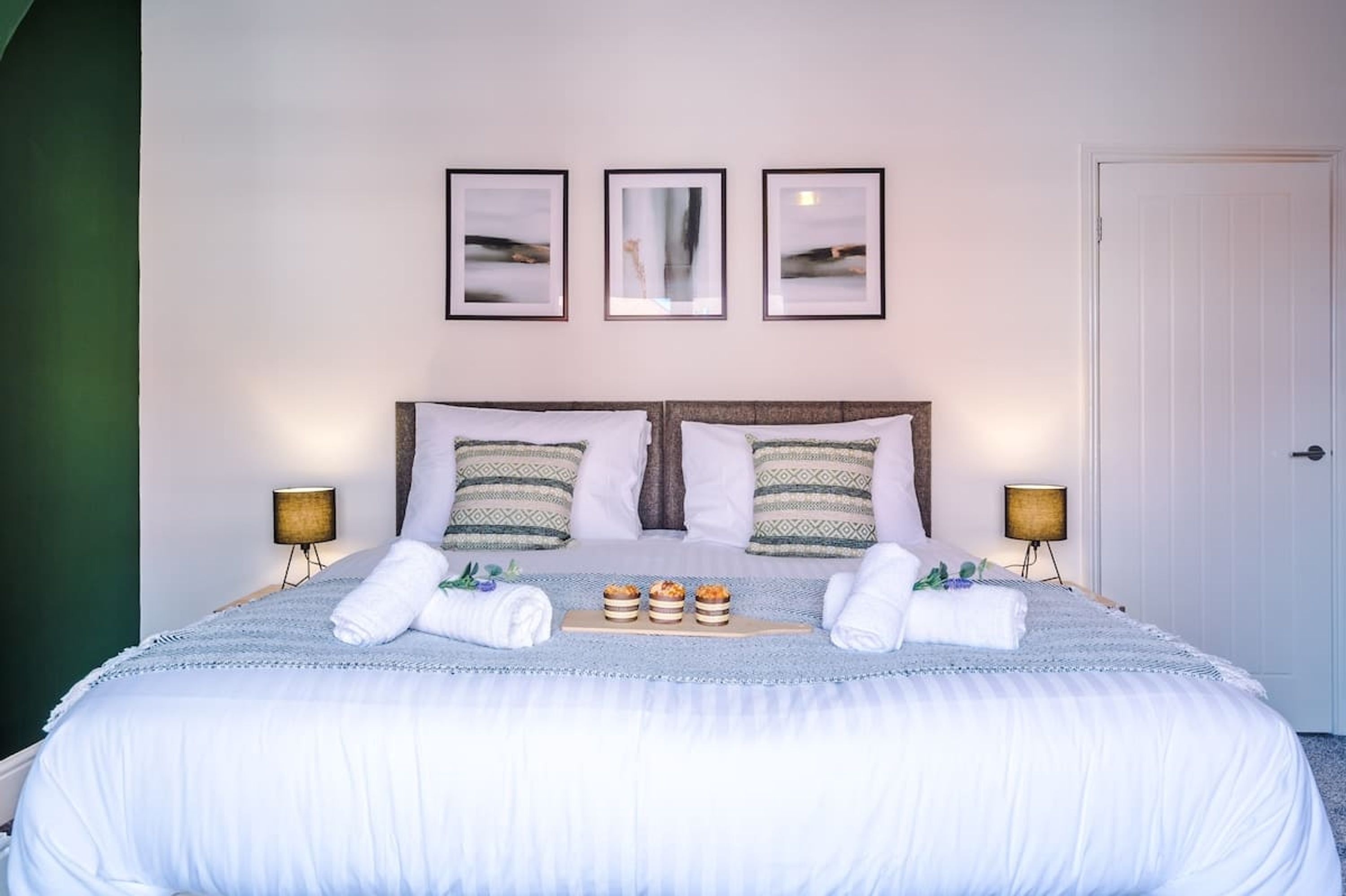 A neatly made bed with a white duvet, two pillows, and a tray of muffins. There are two bedside lamps, a door, and three framed pictures on the wall.