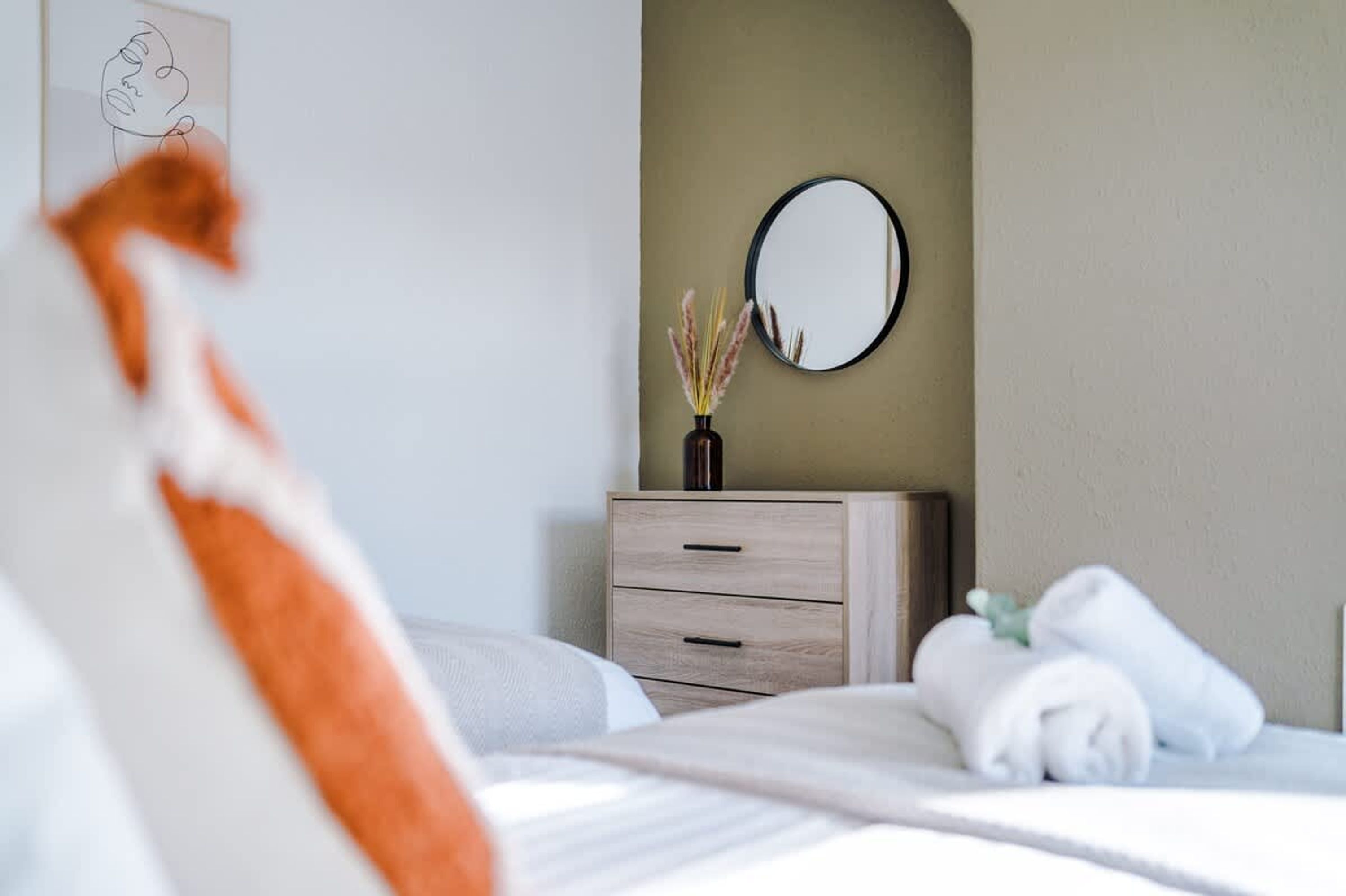 A neatly made bed with white linens, a wooden nightstand with a vase of dried pampas grass, and a round mirror on the wall.