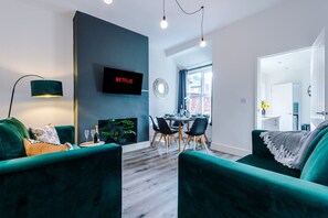 A modern living room with a green sofa, a dining area with a table and chairs, a flat-screen TV mounted on the wall, and a large window. - Modern Home Ideal for Business Groups Sleeps 7 (Crewe)