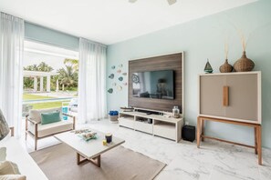A modern living room with a TV, sofa, coffee table, and a view of a pool and greenery. - Two-bedroom apt With Pool View bbq in Lake View (Las Terrenas)