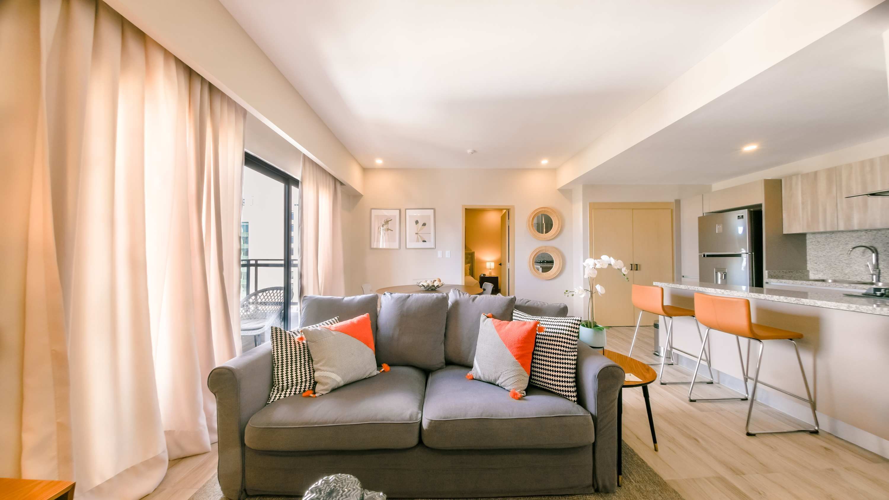Una sala moderna con un sofá gris, cojines estampados en naranja y negro, una zona de comedor con barra y una cocina con electrodomésticos de acero inoxidable.