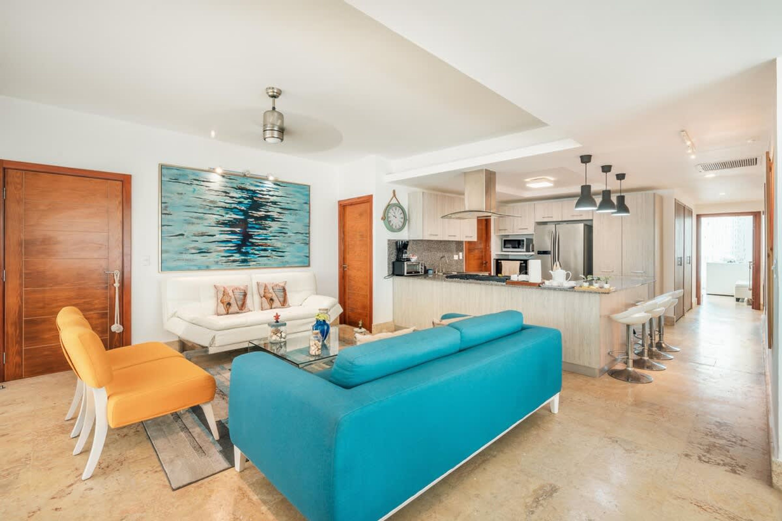 A modern living room with a sofa, a coffee table, and a chair. There is a kitchen area with a refrigerator, microwave, and bar stools.