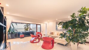 Un salon moderne avec un canapé, des fauteuils rouges, une grande plante et une vue à travers une porte vitrée.