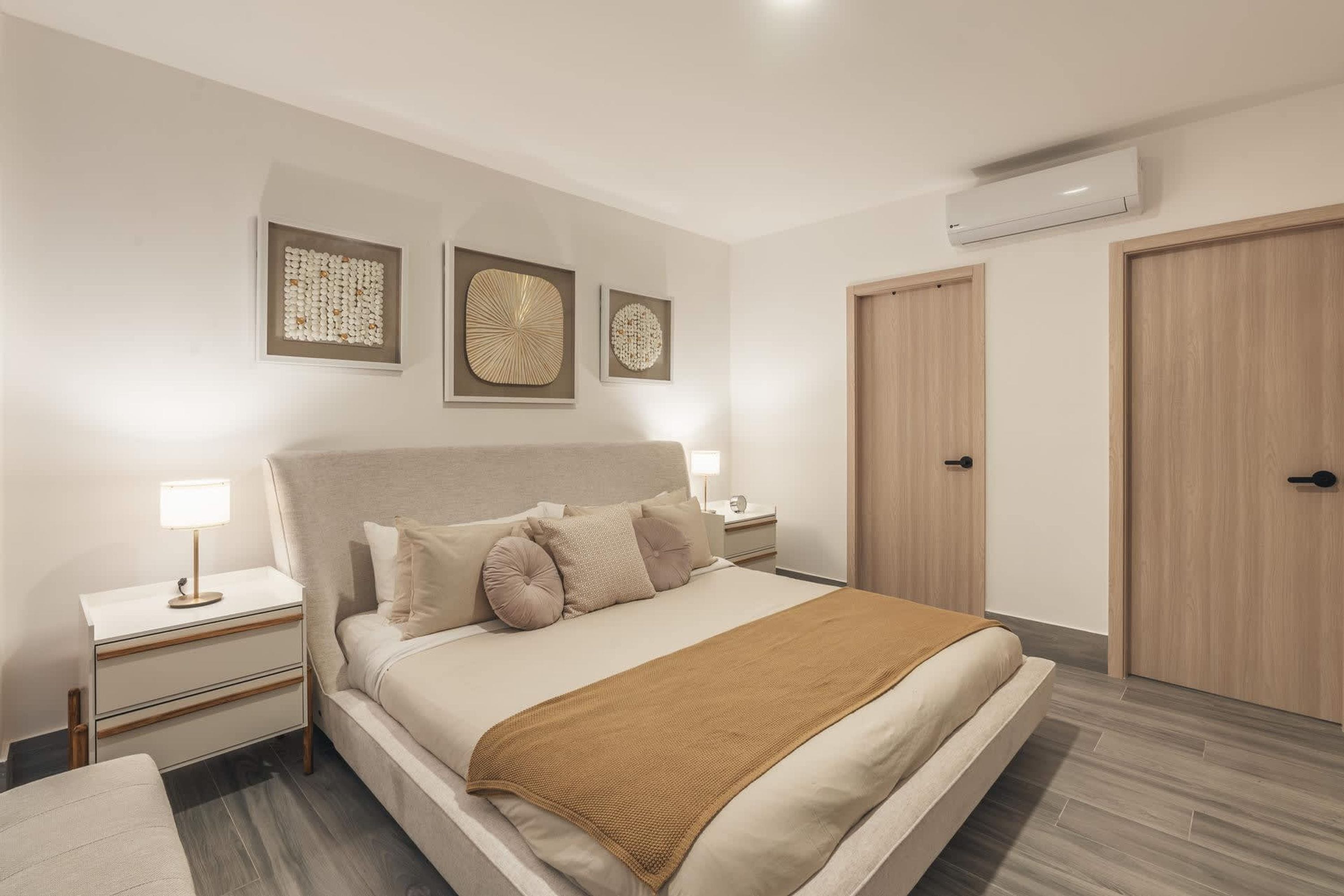 A bedroom with a bed, bedside tables, wall art, an air conditioning unit, and wooden doors.