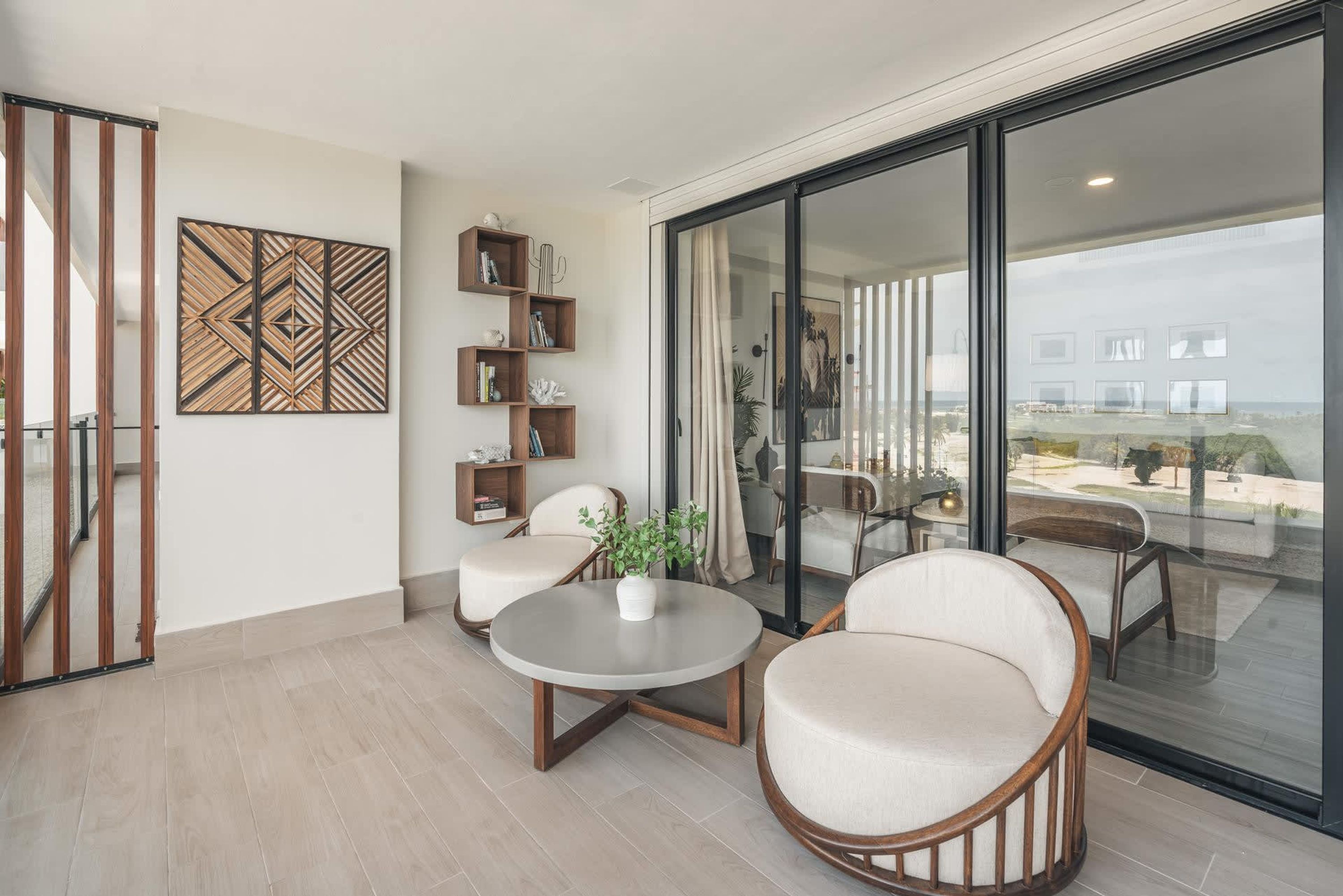 Ein modernes Wohnzimmer mit Glastür zum Balkon, einem runden Couchtisch, zwei gepolsterten Sesseln und einer Wanddekoration.