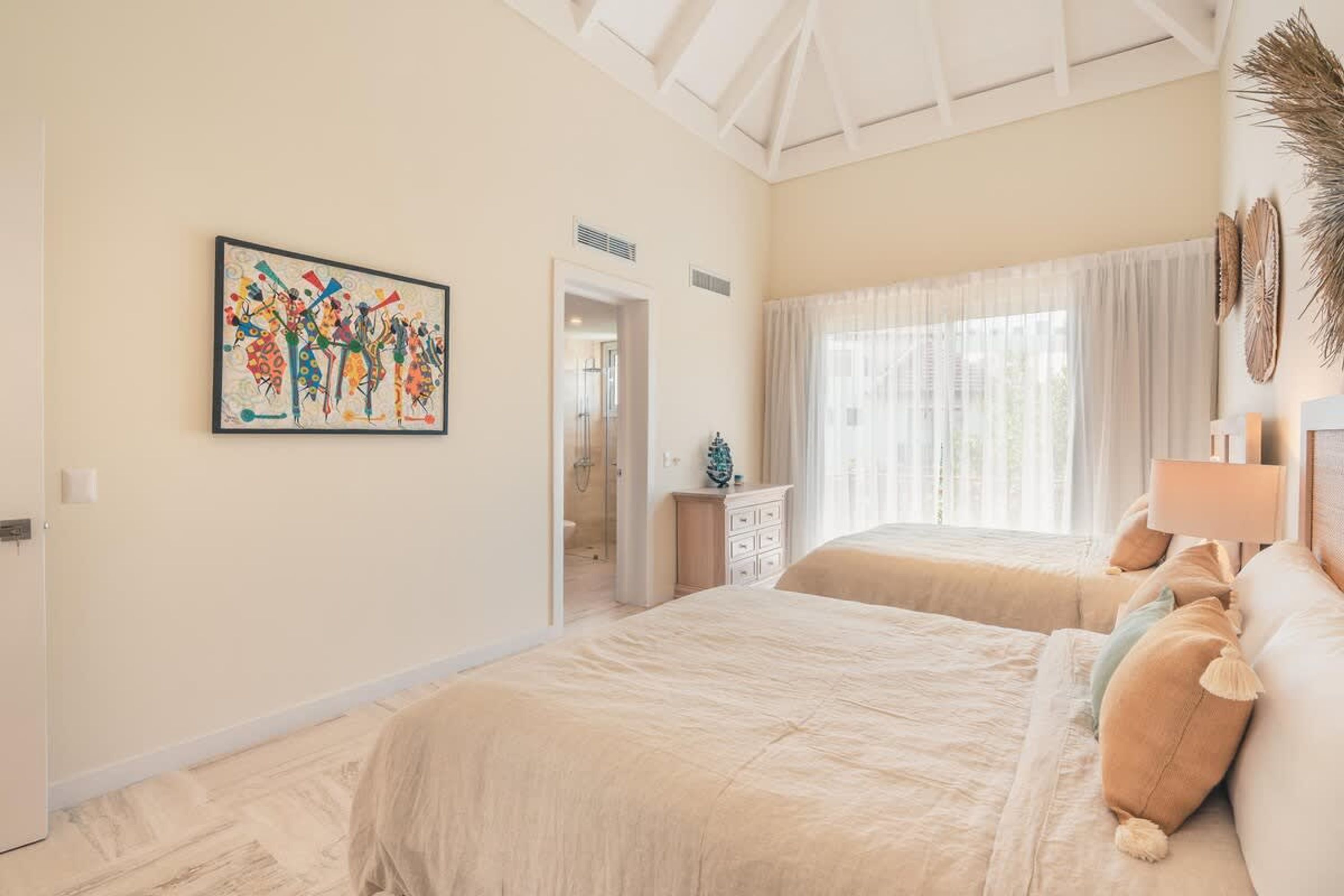 A bedroom with two beds, a large window with sheer curtains, a ceiling fan, and a wall-mounted artwork.