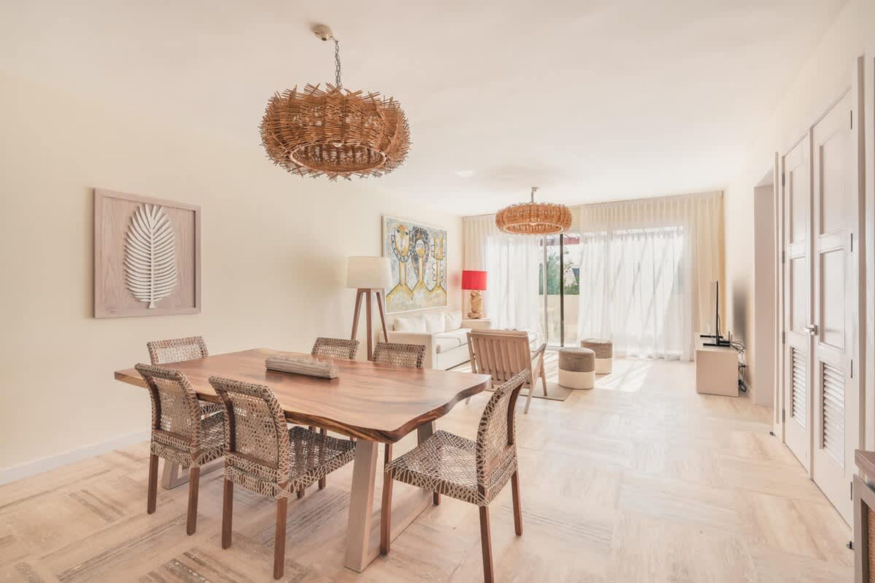 A dining area with a wooden table and wicker chairs, a large woven pendant light, and a painting on the wall.