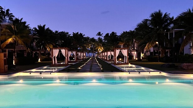 A pool area with a clear view of the water, surrounded by palm trees and a row of wooden gazebos. - 2br Apt 5 Guests Balcony With Pool View (Las Terrenas)