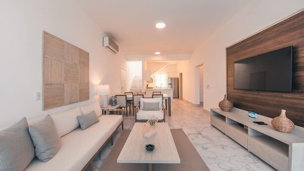 A modern living room with a sofa, coffee table, TV, and a kitchen in the background. - 3br-penthouse w Jacuzzi Incl at Lake View (Las Terrenas)