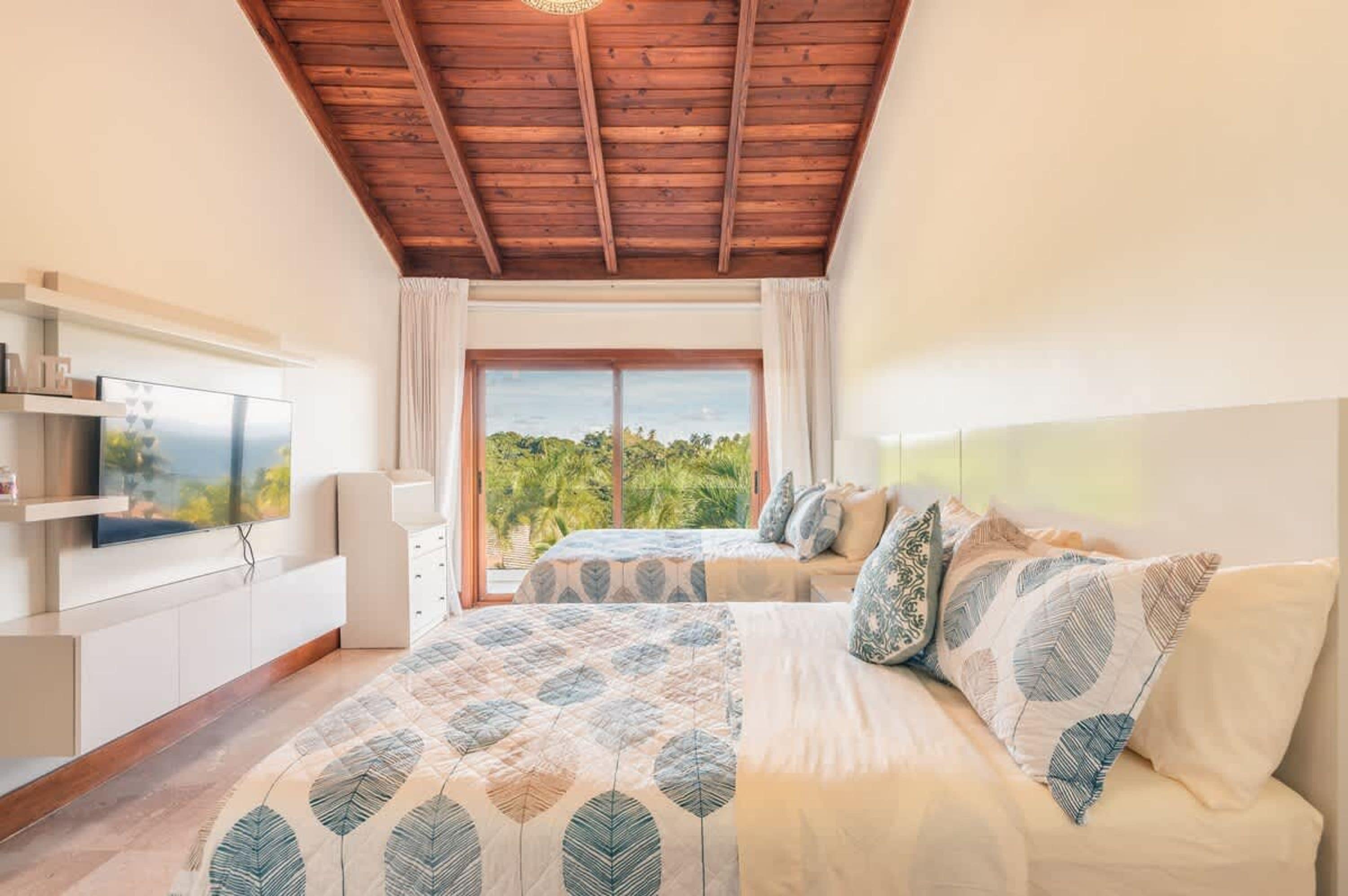 A bedroom with a large bed, a TV, a wooden ceiling, and a view of greenery.