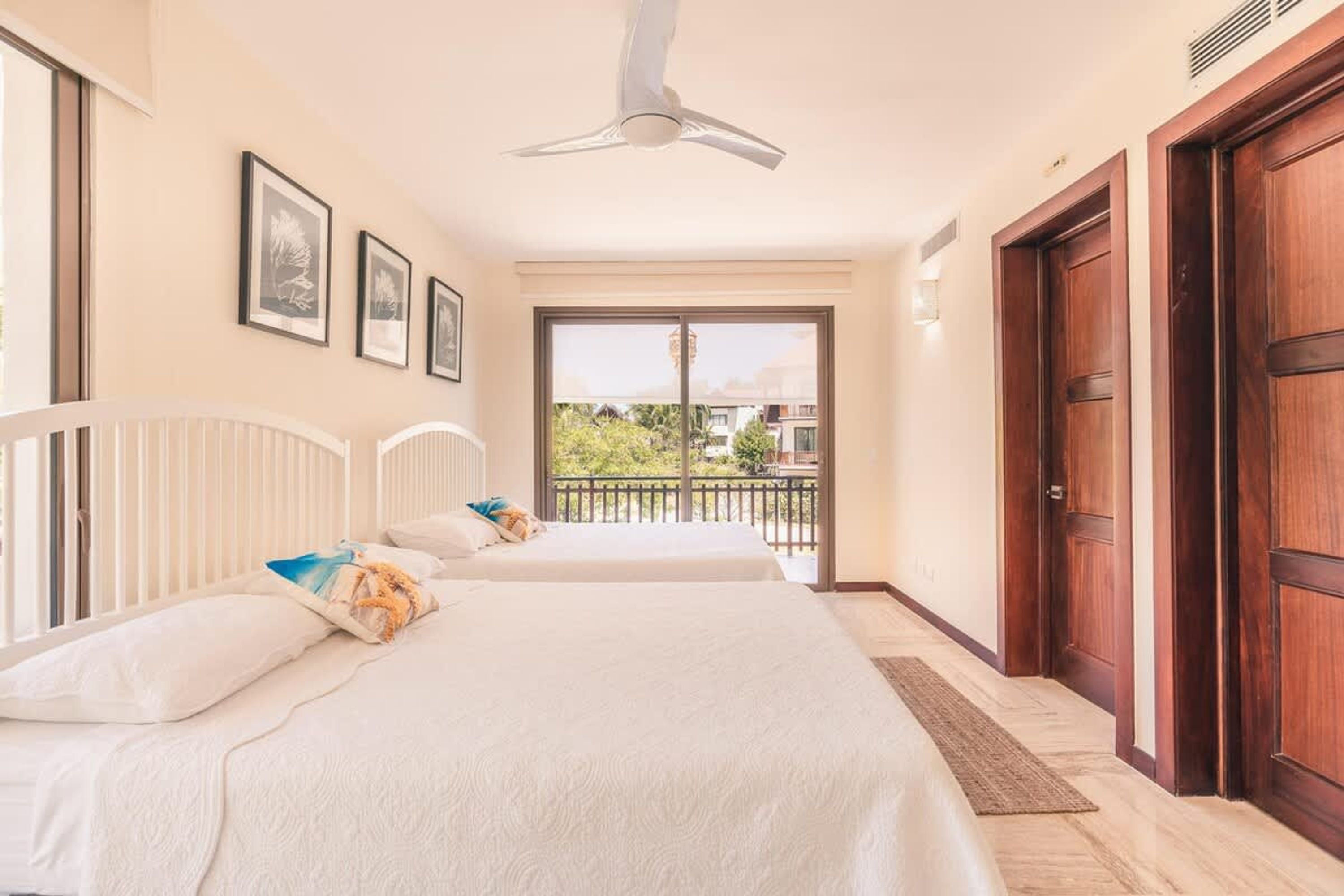 A bedroom with two beds, a ceiling fan, a balcony with a view, and wooden doors.