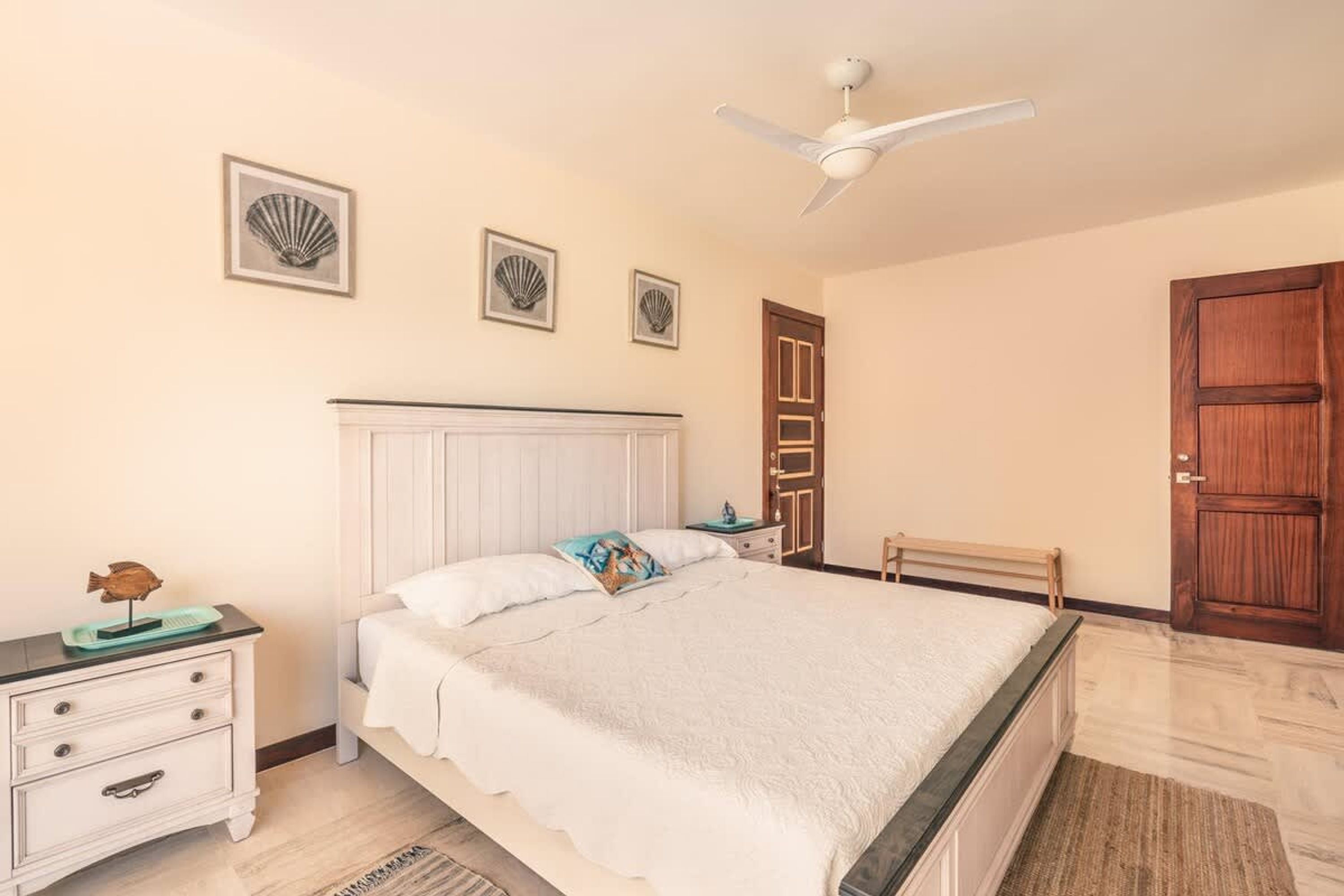 Une chambre à coucher comprenant un lit, une table de chevet, une commode, un ventilateur de plafond et des tableaux encadrés accrochés au mur.