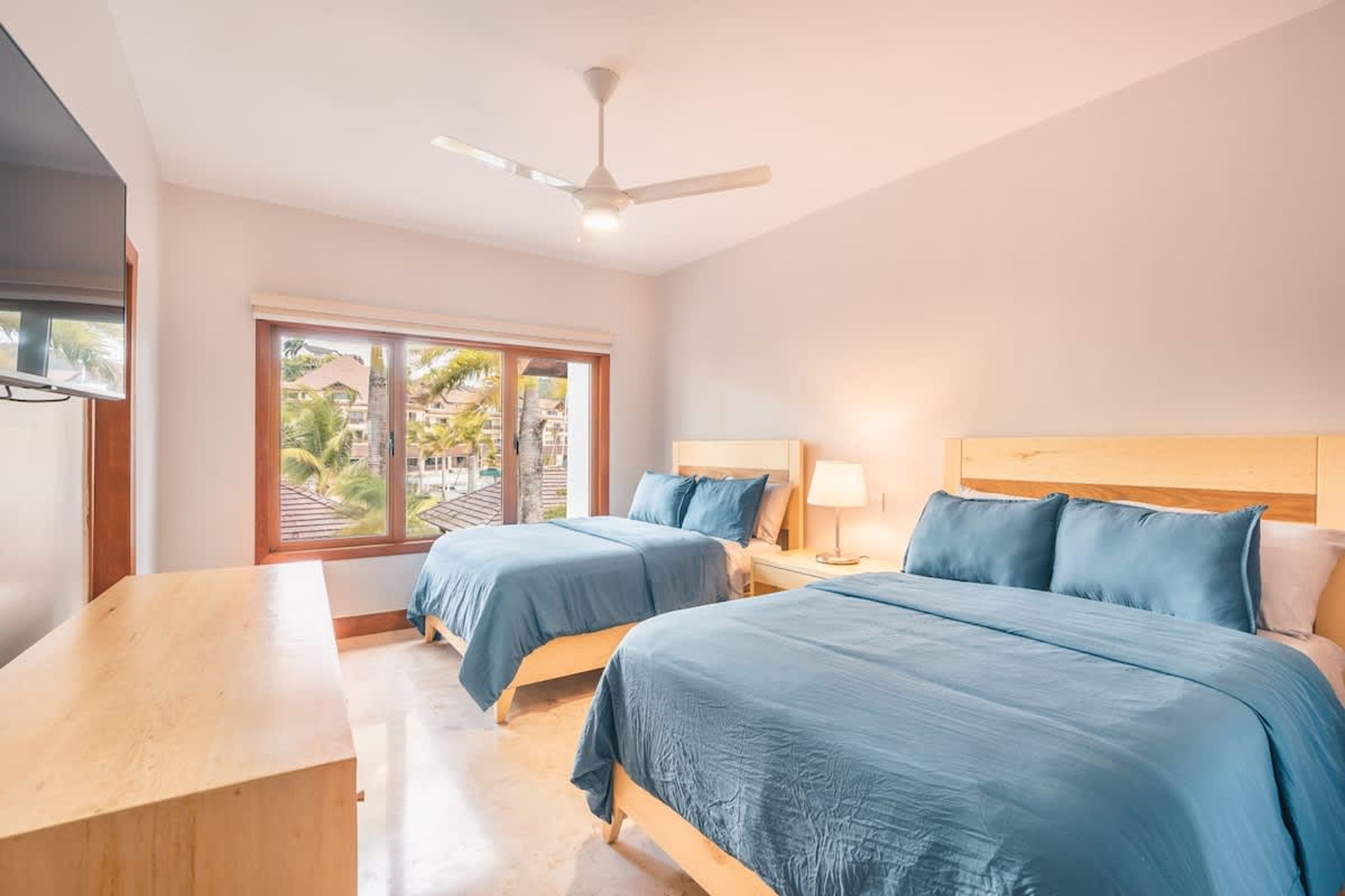 Habitación de hotel con dos camas, un escritorio, ventilador de techo y una ventana grande con vistas a palmeras.