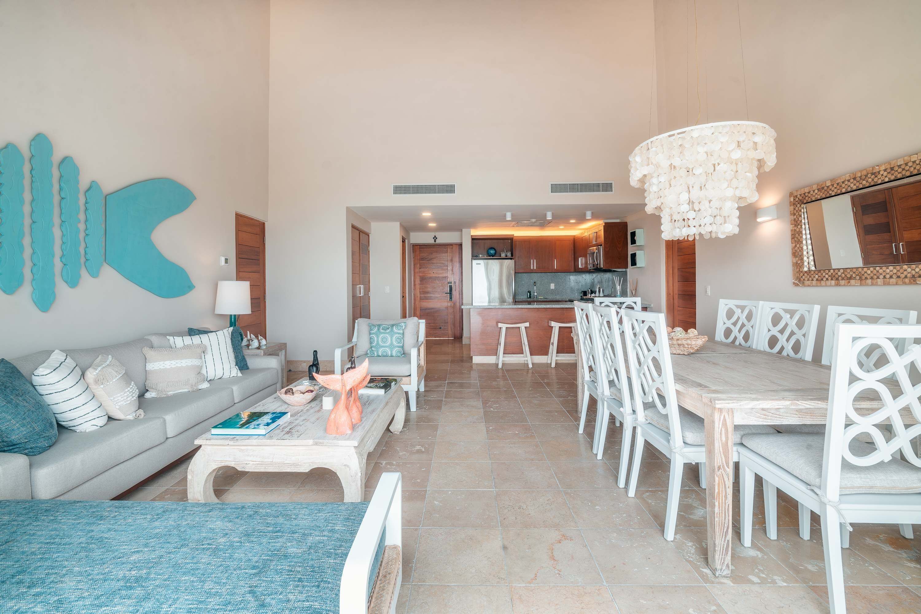 A spacious living area with a sofa, coffee table, dining table with chairs, a kitchen in the background, and a chandelier.