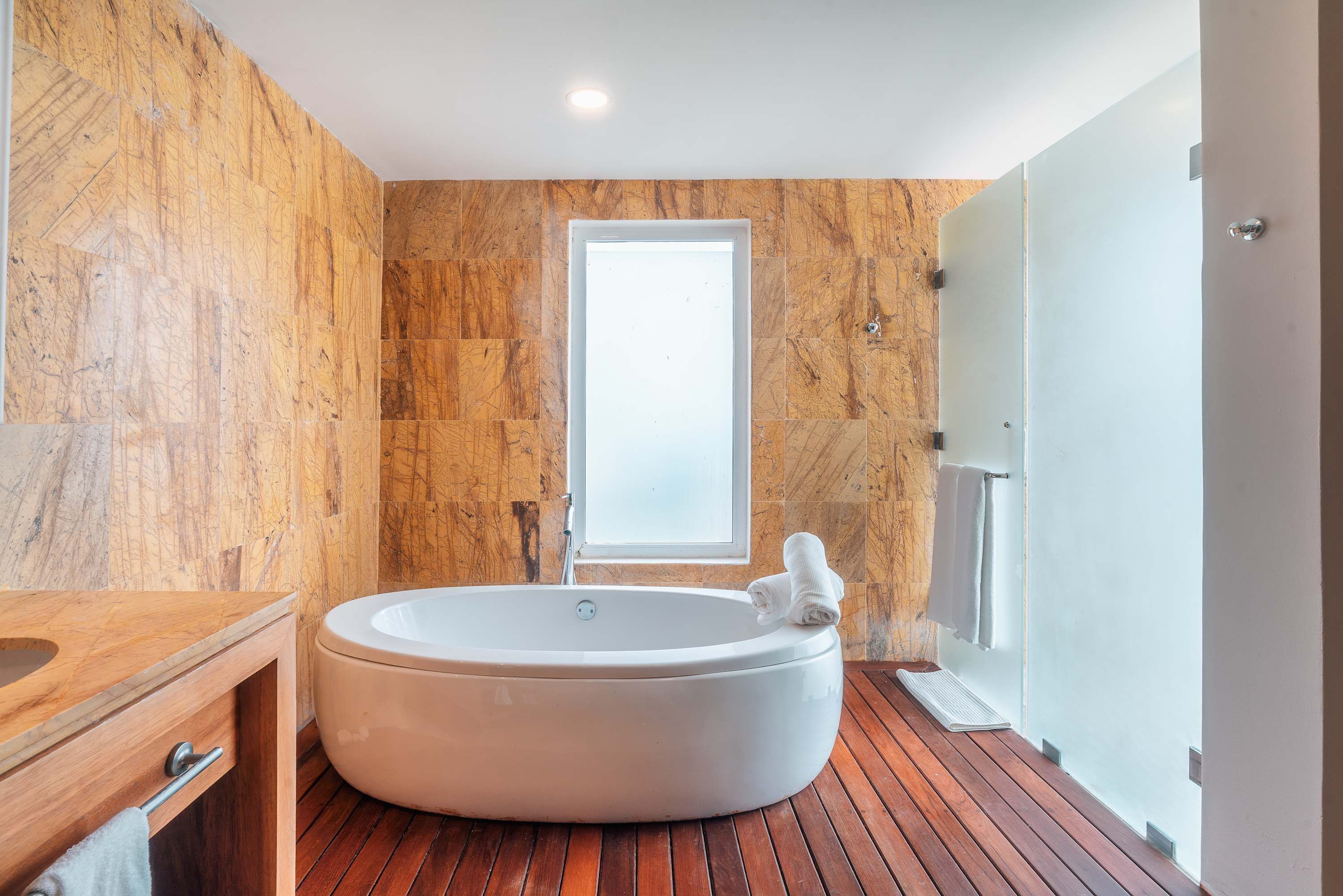A modern bathroom with a large bathtub, wooden flooring, a wooden vanity, and a large window.