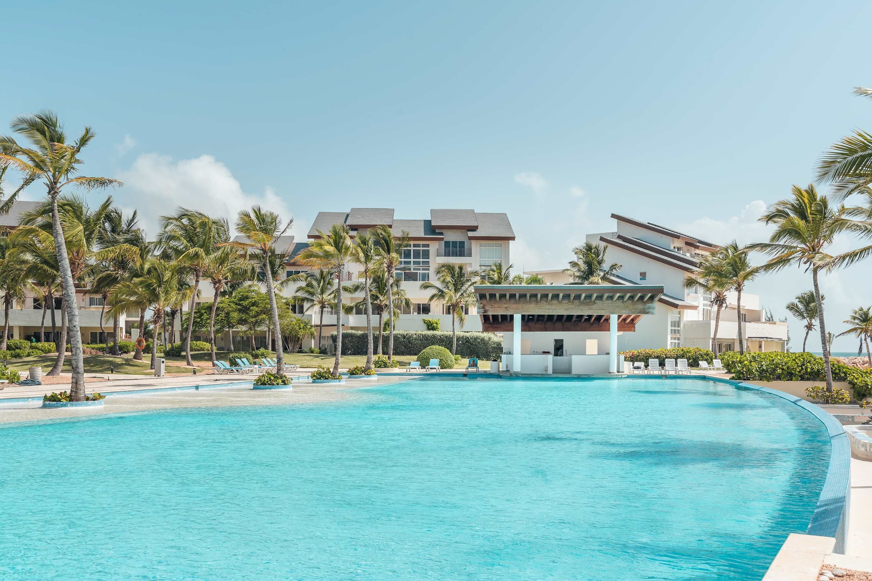 Piscina de resort con palmeras, un área techada y edificios modernos al fondo.