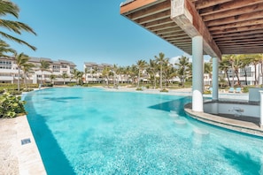 A clear swimming pool with a wooden pergola overhead, surrounded by palm trees and hotel buildings.