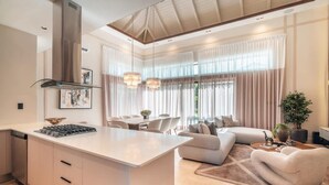 Cocina moderna con una isla central, comedor con mesa y sillas, un sofá y un amplio ventanal con cortinas.