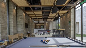 Mesa de billar con taco, mesa de futbolín y zonas de estar en un espacio interior moderno con paredes de ladrillo y ventanas grandes.