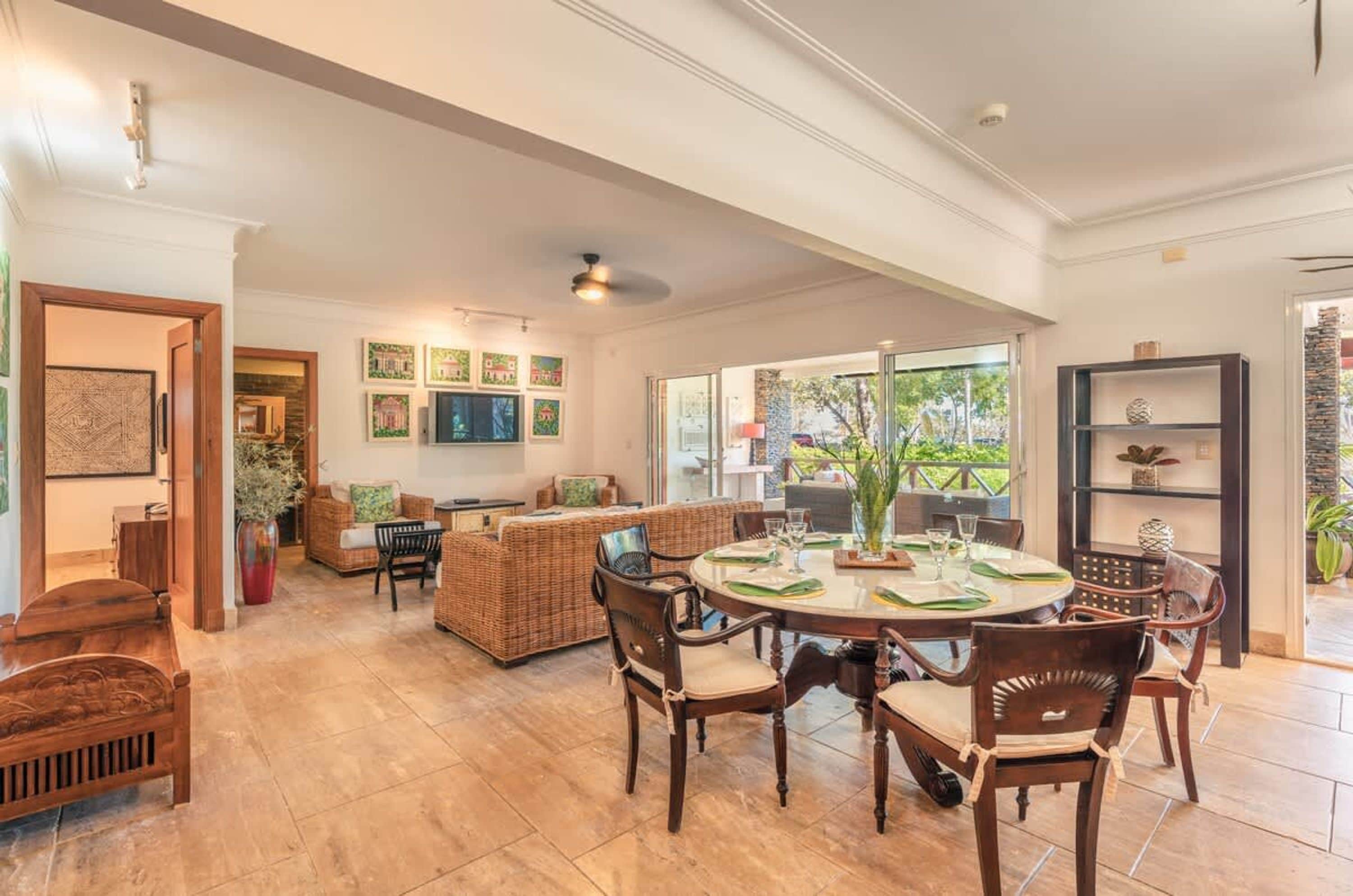 A spacious living area with a round dining table, wicker furniture, and a large window offering a view of the outdoors.