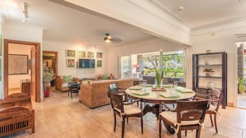 A spacious living area with a round dining table, wicker furniture, and a large window offering a view of the outdoors.