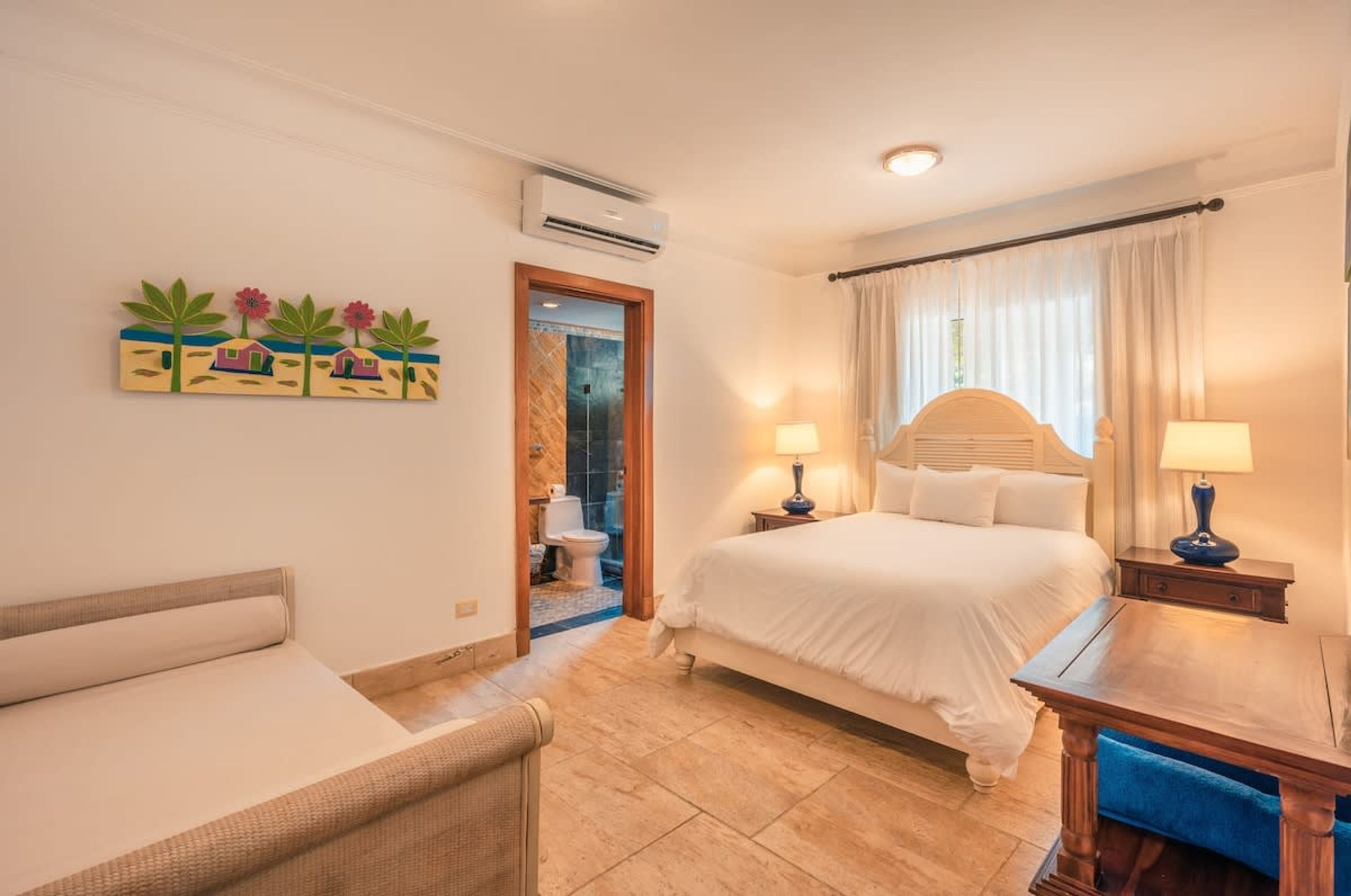 A hotel room with a bed, two bedside tables with lamps, a chair, a painting on the wall, and an air conditioning unit.