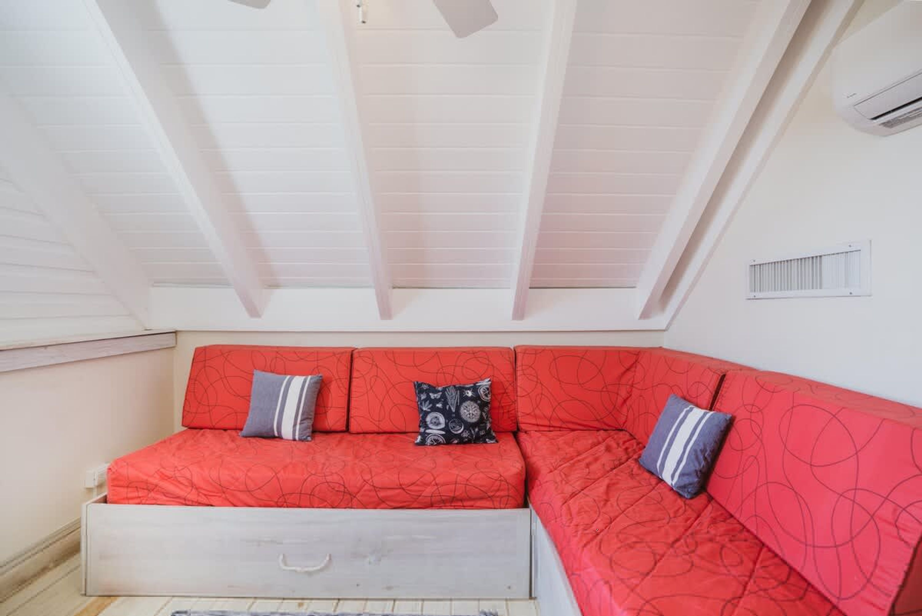 Un canapé-lit rouge agrémenté de coussins à motifs, un plafond blanc avec des poutres apparentes et une petite fenêtre dotée d’une grille d’aération.
