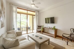 A modern living room with a sofa, coffee table, and chairs, a TV mounted on the wall, and a sliding glass door leading to an outdoor area. - Elegant 1br Casa de Campo Retreat w Garden Views (La Romana)