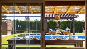A view through a window of a patio area with a pool, outdoor furniture, and a wooden pergola. - Sunnyvalley by Fidalsa (Dolores)