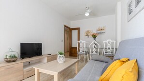 A modern living room with a grey sofa, a wooden coffee table, a TV on a wooden stand, and a dining area with white chairs and a table. - Fidalsa Bay View Romance (Guardamar del Segura)