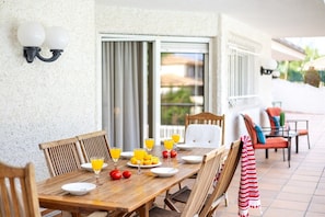 A wooden dining table set with plates, glasses, and fruits on a patio with chairs and a striped towel.
