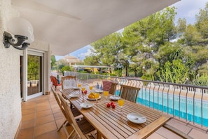 A wooden table set with plates, glasses, and fruit on a balcony with a pool view.