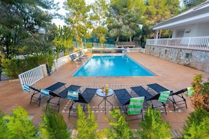 A rectangular swimming pool with a tiled deck, surrounded by a white fence. There are lounge chairs and a table with a bottle on it.