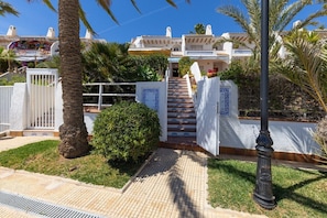 Un edifici residencial amb una escala que condueix a una entrada tancada per reixes, envoltat de palmeres i un jardí ben cuidat.