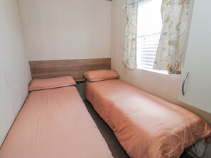 Un petit logement composé d’une seule pièce, comprenant deux lits, une tête de lit en bois, une fenêtre avec des stores et une poignée de porte.