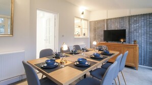 A dining area with a long table set for six, chairs, a television, and a kitchen area in the background. - Beau Jardin (Telford)