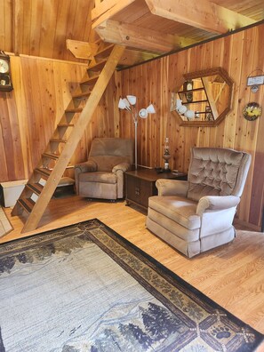 A wooden interior with a staircase, two armchairs, a lamp, and a rug.