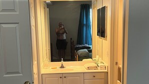 Une petite chambre à coucher, de taille réduite, comprenant un lit simple, une table de chevet, un miroir et une fenêtre avec des rideaux.