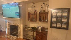 Un salon avec une cheminée, un téléviseur diffusant un match de golf, des clubs de golf rangés sur des étagères et une carte encadrée accrochée au mur.