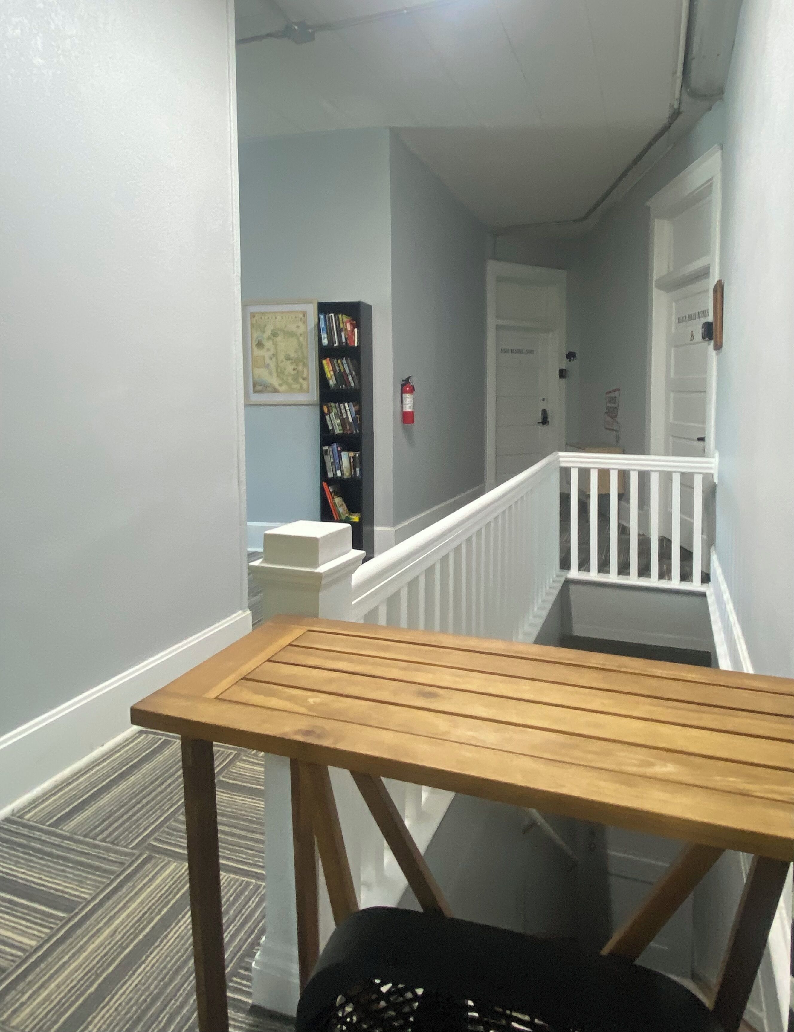 A hallway with a wooden table, a black chair, a bookshelf, and a white railing.