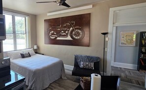 A bedroom with a bed, a ceiling fan, a wall-mounted motorcycle artwork, a chair with a towel, a bookshelf, and a door with a map. - Iron Rider Studio - Couples Motorcycle Theme Loft Walkable Downtown Hot Springs (Hot Springs)
