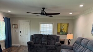 Uma sala de estar com ventilador de teto, um sofá cinza, um banquinho estampado, um tapete cinza e piso de madeira.