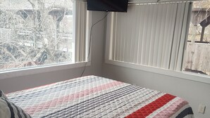 Un dormitorio con cama, televisor montado en la pared y ventana con persianas.