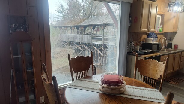 Una mesa de comedor de madera con sillas, una ventana que ofrece vistas a una construcción de madera en el exterior, y una zona de cocina con armarios y electrodomésticos.