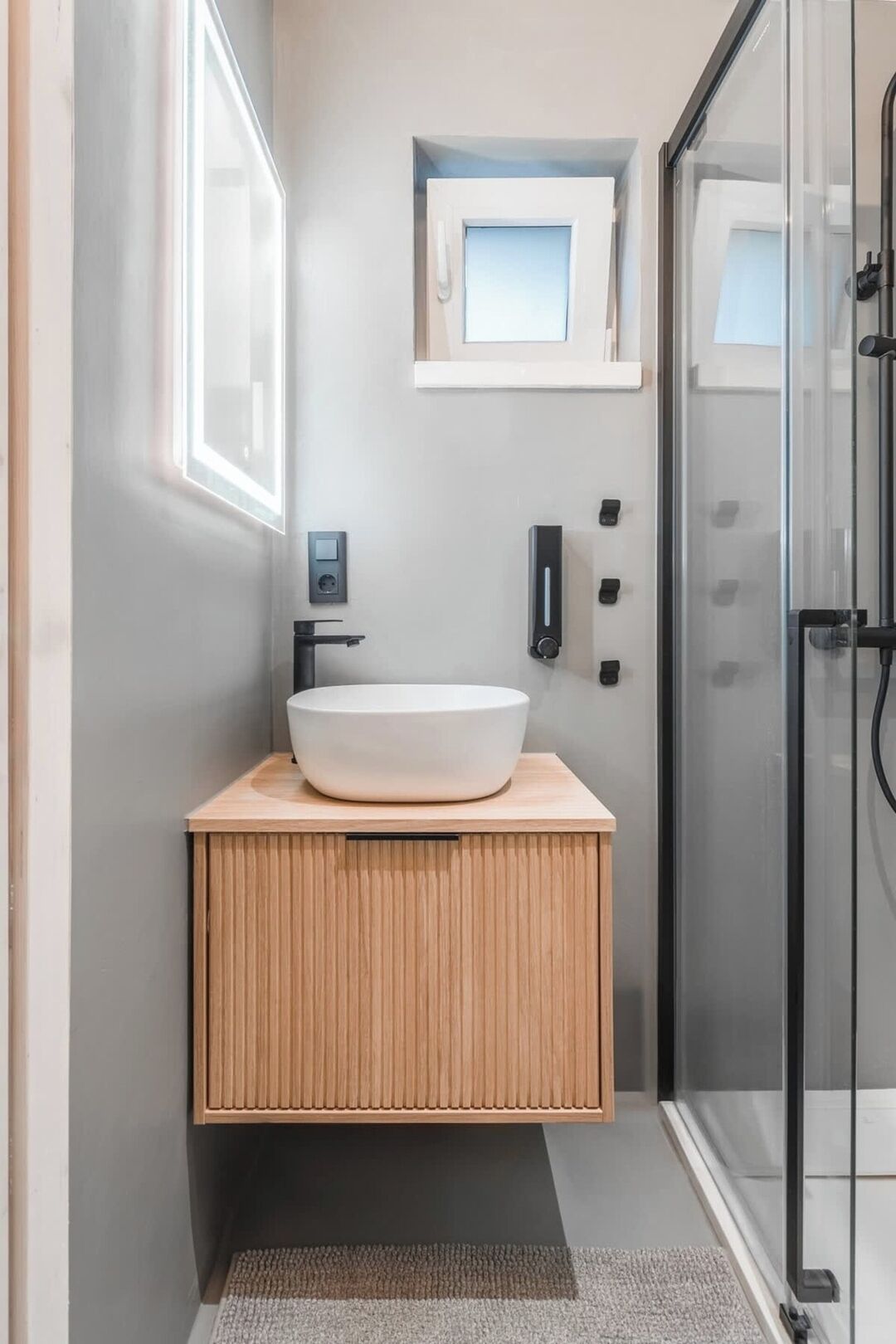 Une salle de bain moderne avec un grand lavabo blanc, un meuble-lavabo en bois, une cabine de douche en verre et une petite fenêtre au-dessus du miroir.