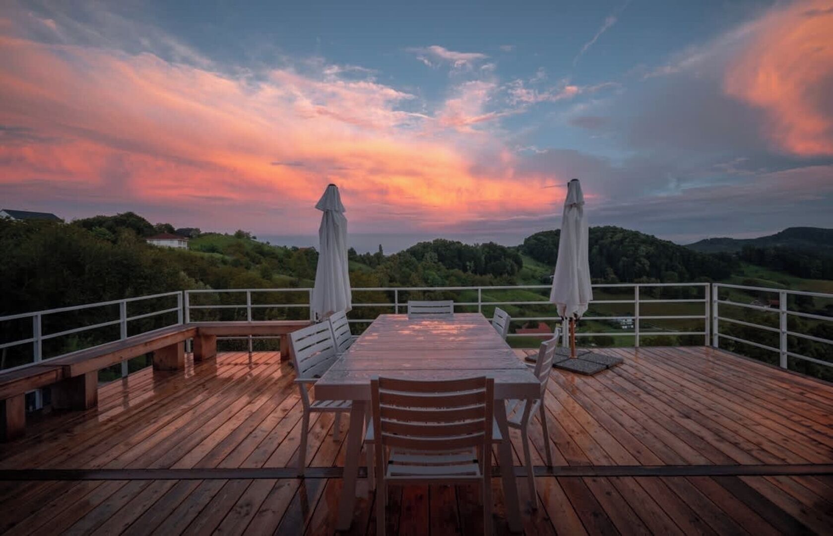 Een houten terras met een tafel, stoelen en twee parasols, met uitzicht op het landschap.